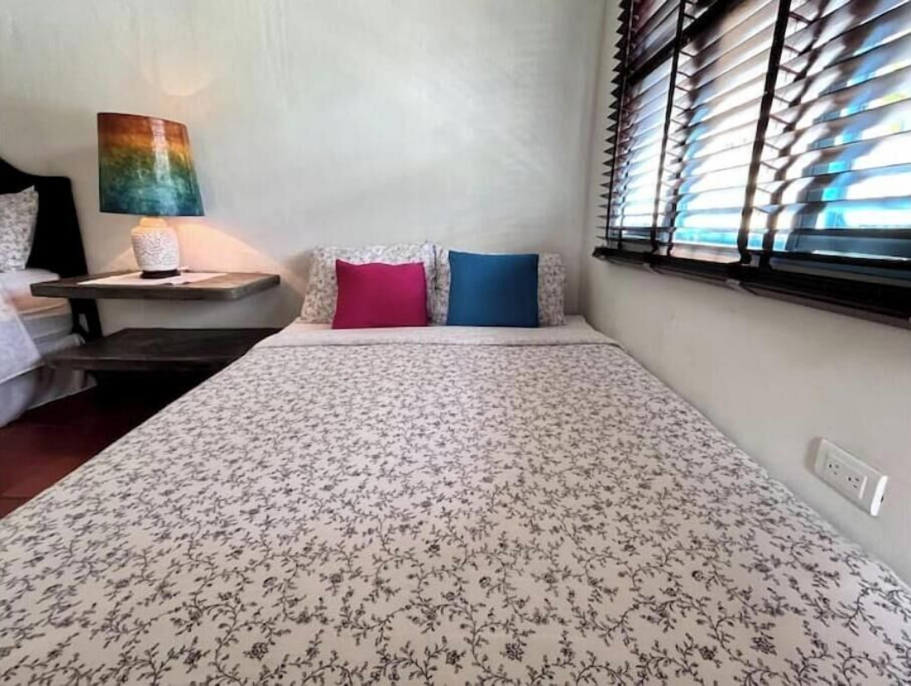 Een slaapkamer met een bloemenprint bedsprei, een nachtkastje met een lamp, en een raam met jaloezieën.