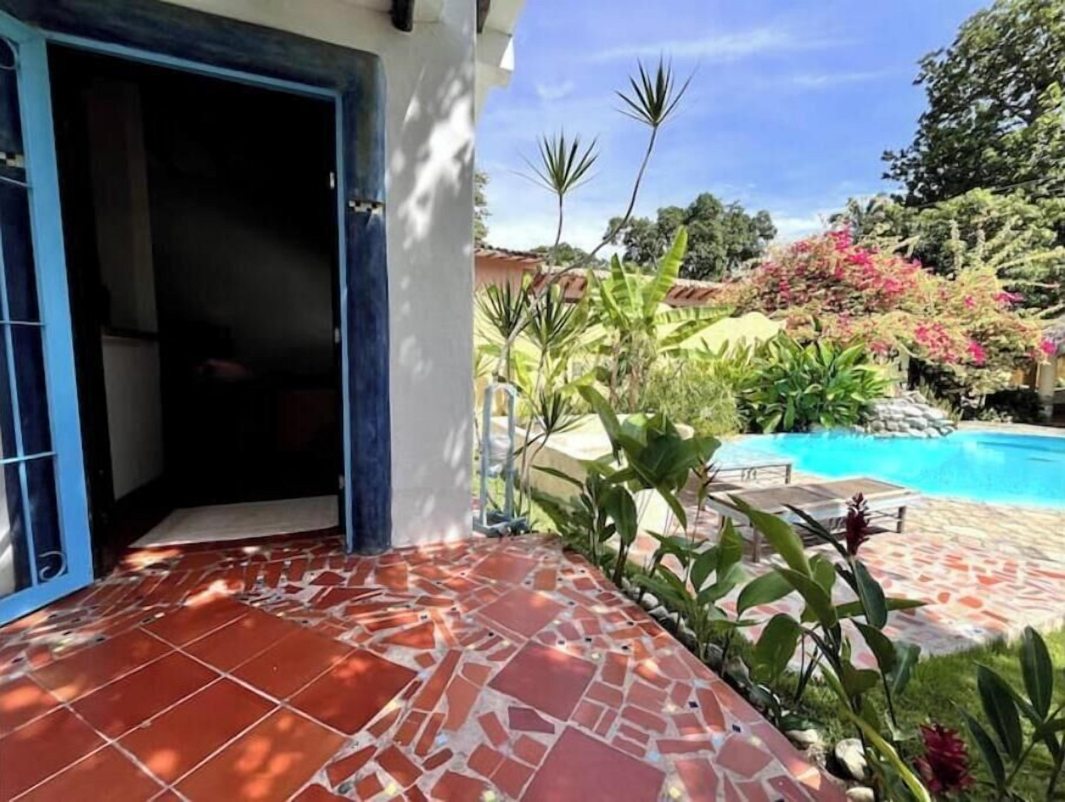 Un pati amb rajoles i una piscina, envoltat de vegetació abundant i un cel blau clar.