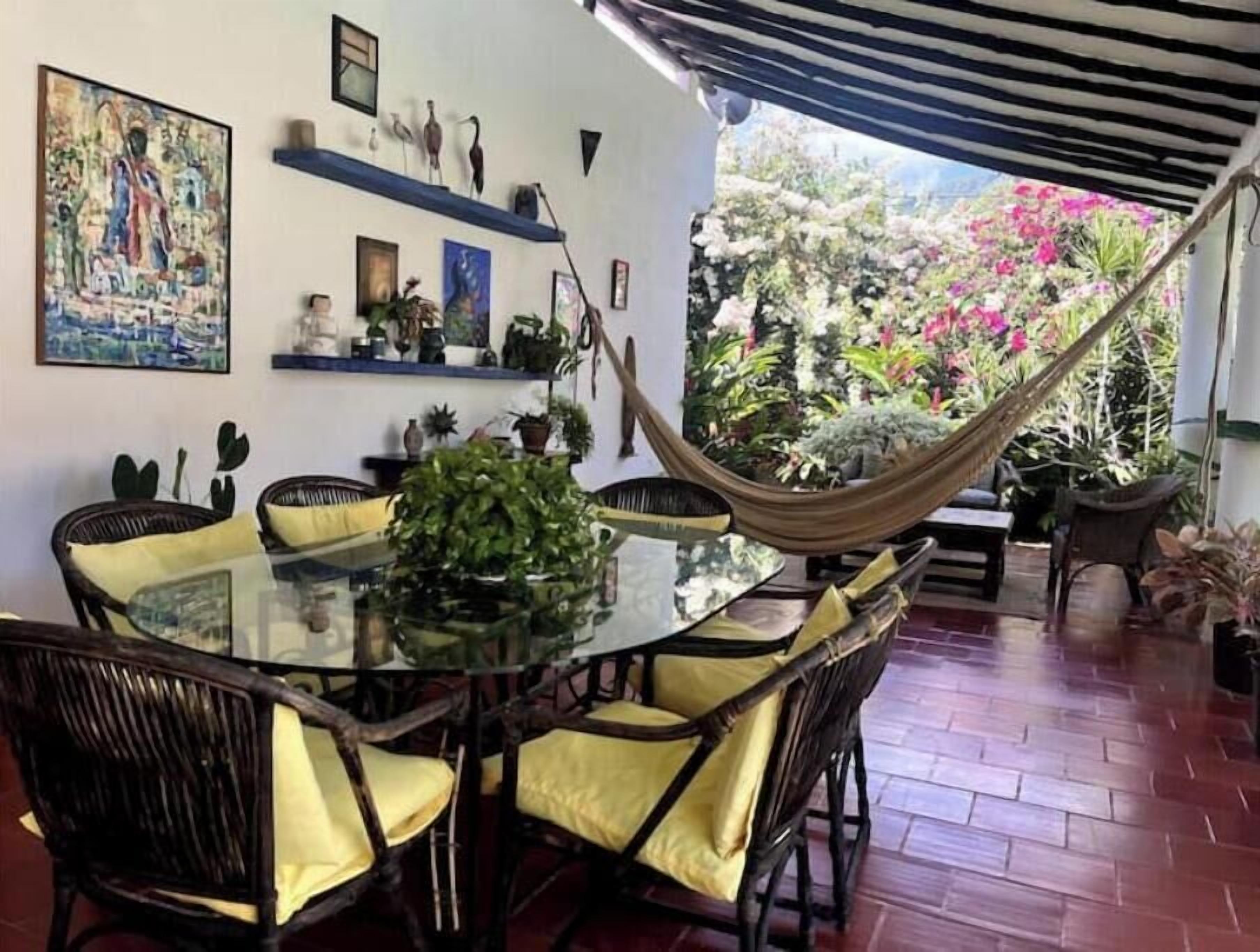 Een eetruimte met een glazen tafel, omringd door rieten stoelen, een hangmat en uitzicht op een tuin met roze bloemen.