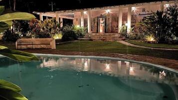 Une vue nocturne d’un espace piscine, avec un bâtiment bien éclairé en arrière-plan, entouré d’une végétation luxuriante.