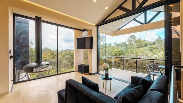 Una sala de estar moderna con un sofá grande, un televisor montado en la pared y un balcón con vistas al entorno verde.
