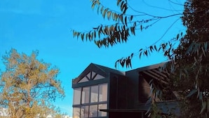 A modern house with a large glass window section, surrounded by trees and a clear blue sky.