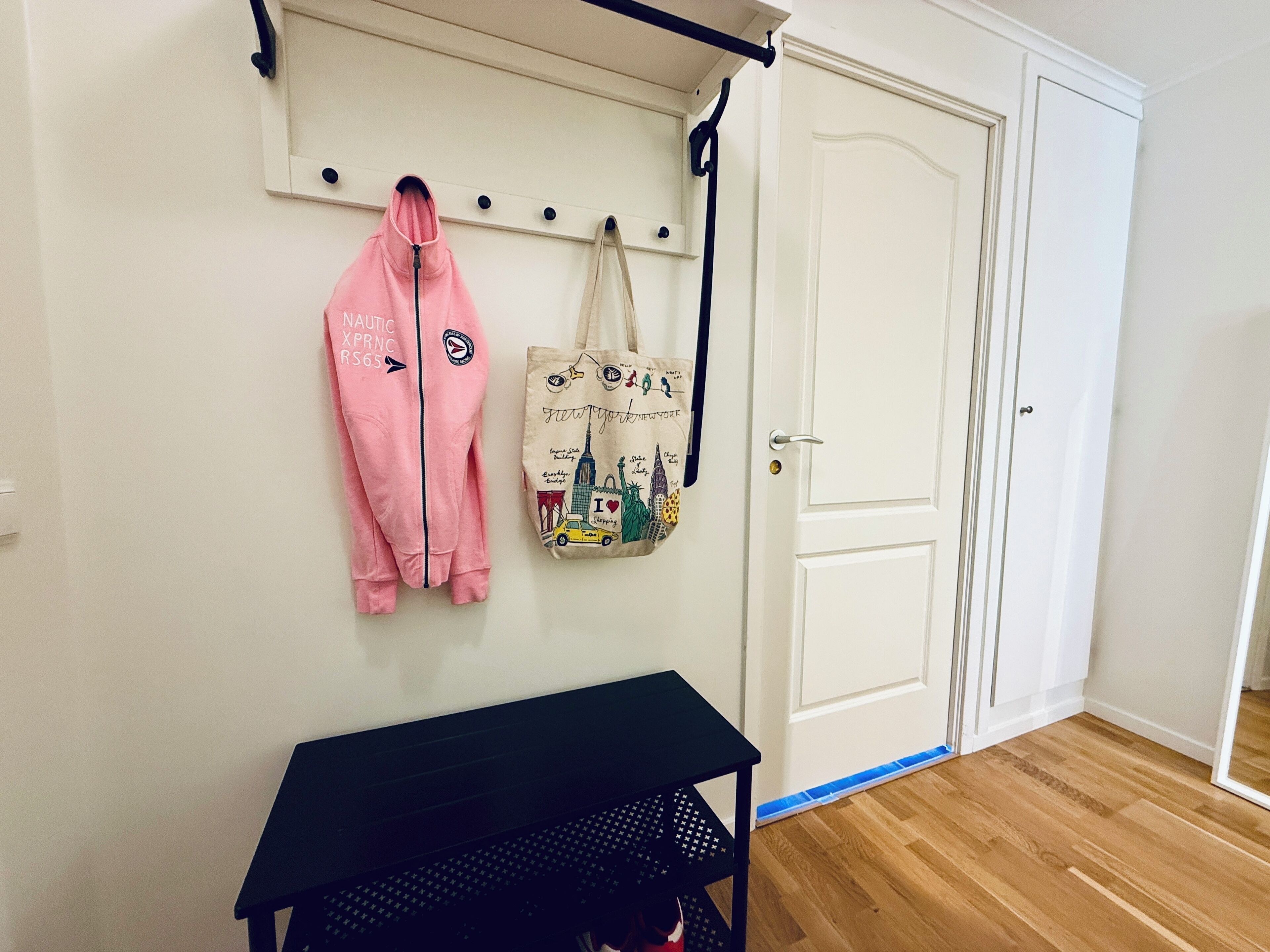 Une pièce avec une porte blanche, une table noire et un porte-manteau mural sur lequel sont accrochés un blouson rose et un sac fourre-tout.