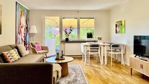 A living room with a sofa, coffee table, dining table, and chairs. There is a TV, a painting, and a window with a view of greenery. - Brilliant family getaway in Stockholm - 4 Guests (Kista)