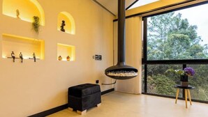 Un intérieur moderne avec un pouf noir, des étagères ornées d’objets décoratifs, une cheminée suspendue et une fenêtre donnant sur les arbres.