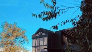 Une maison moderne dotée d’une grande baie vitrée, entourée d’arbres et surplombant un ciel bleu azur.