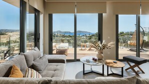 A modern living room with a large sofa, a coffee table, and a view of a deck with outdoor furniture. - Villa Elounda Lion, 2 Pools, Hammam & Close to Sea (Agios Nikolaos)