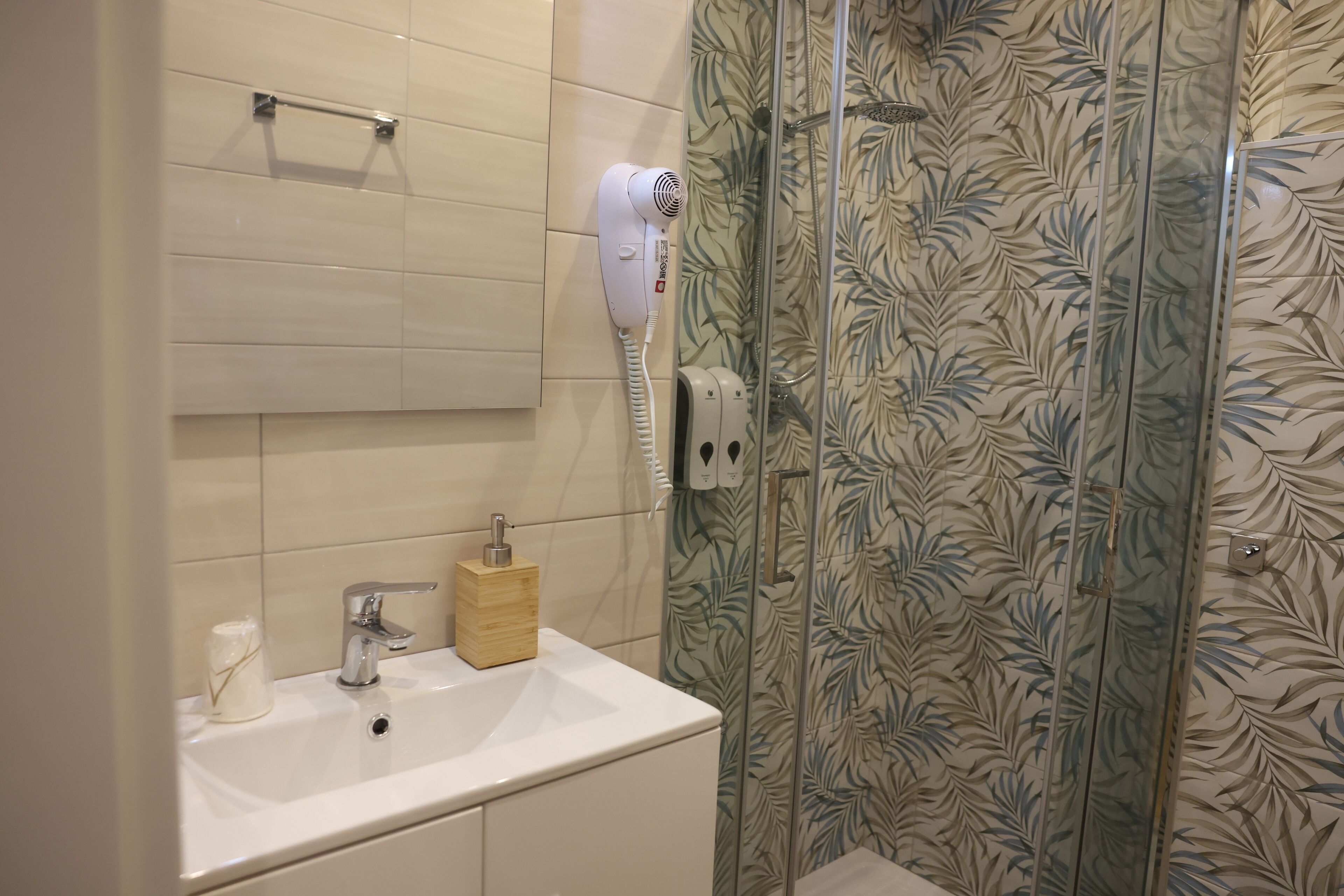 A bathroom with a white sink, a hairdryer on the wall, and a shower with a leaf-patterned shower curtain.