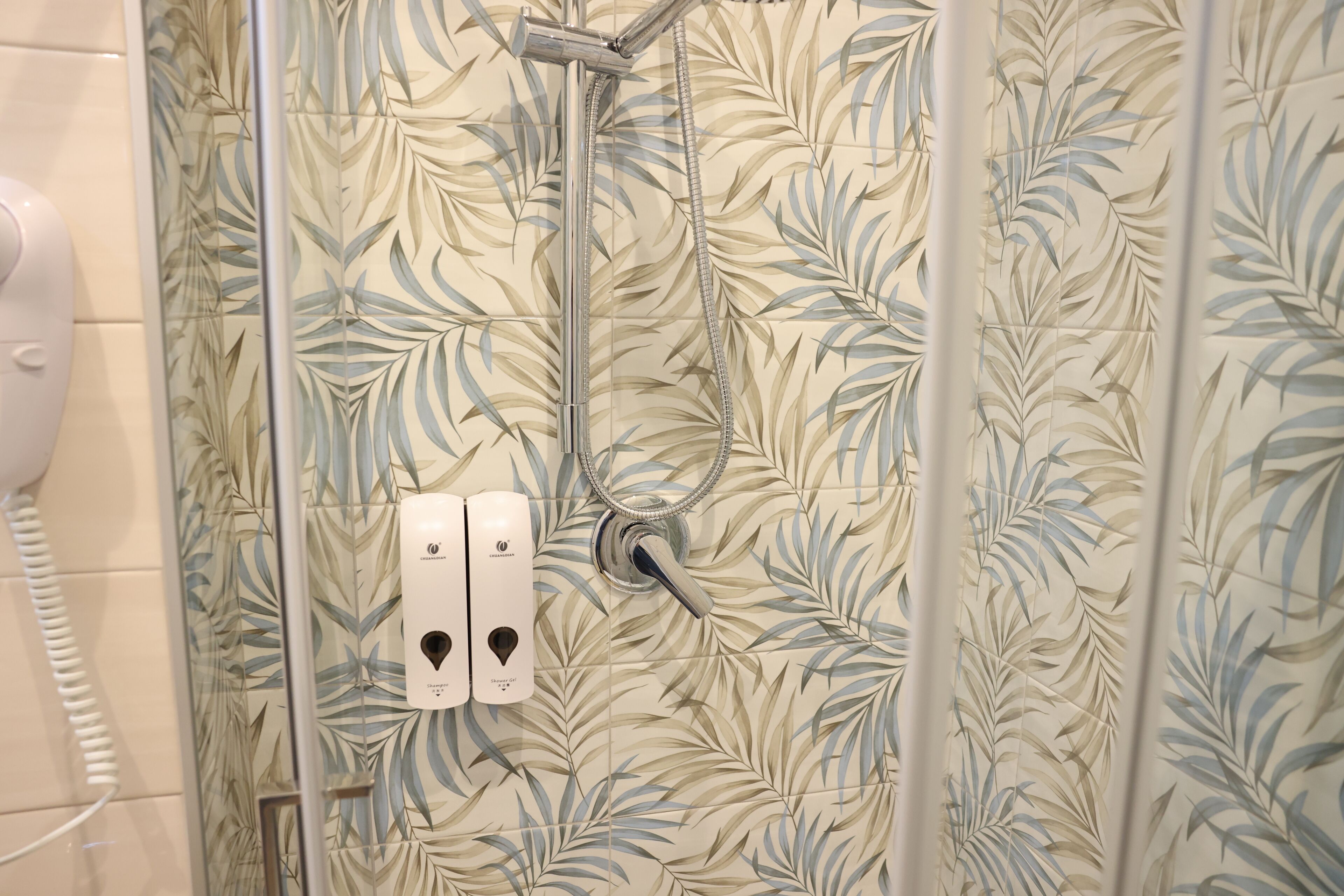 Ducha con pared de azulejos con diseño de hojas, cabezal de ducha manual y secamanos blanco.