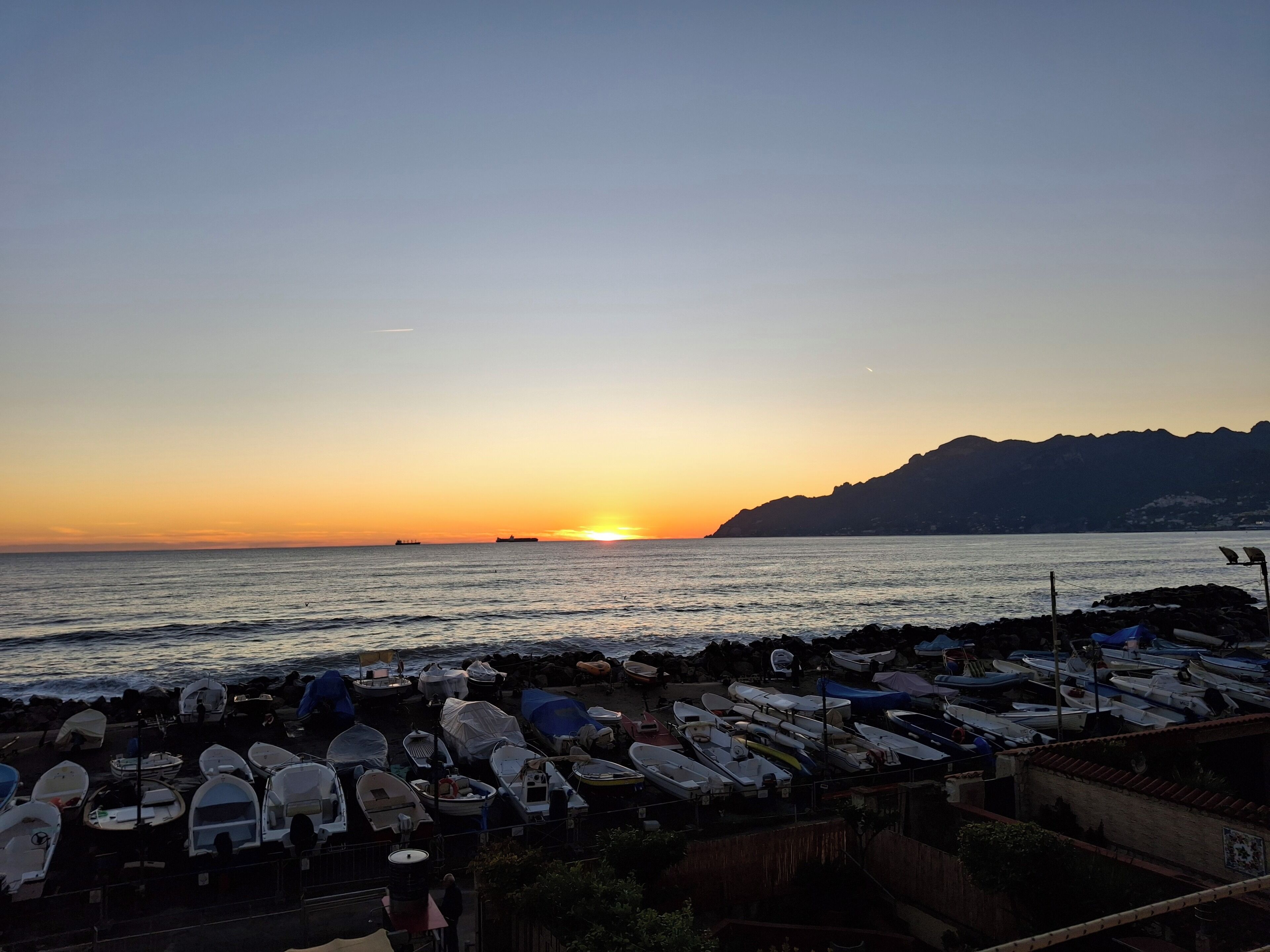 Un paysage côtier avec des bateaux amarrés le long de la rive, un coucher de soleil en arrière-plan et un paysage montagneux.
