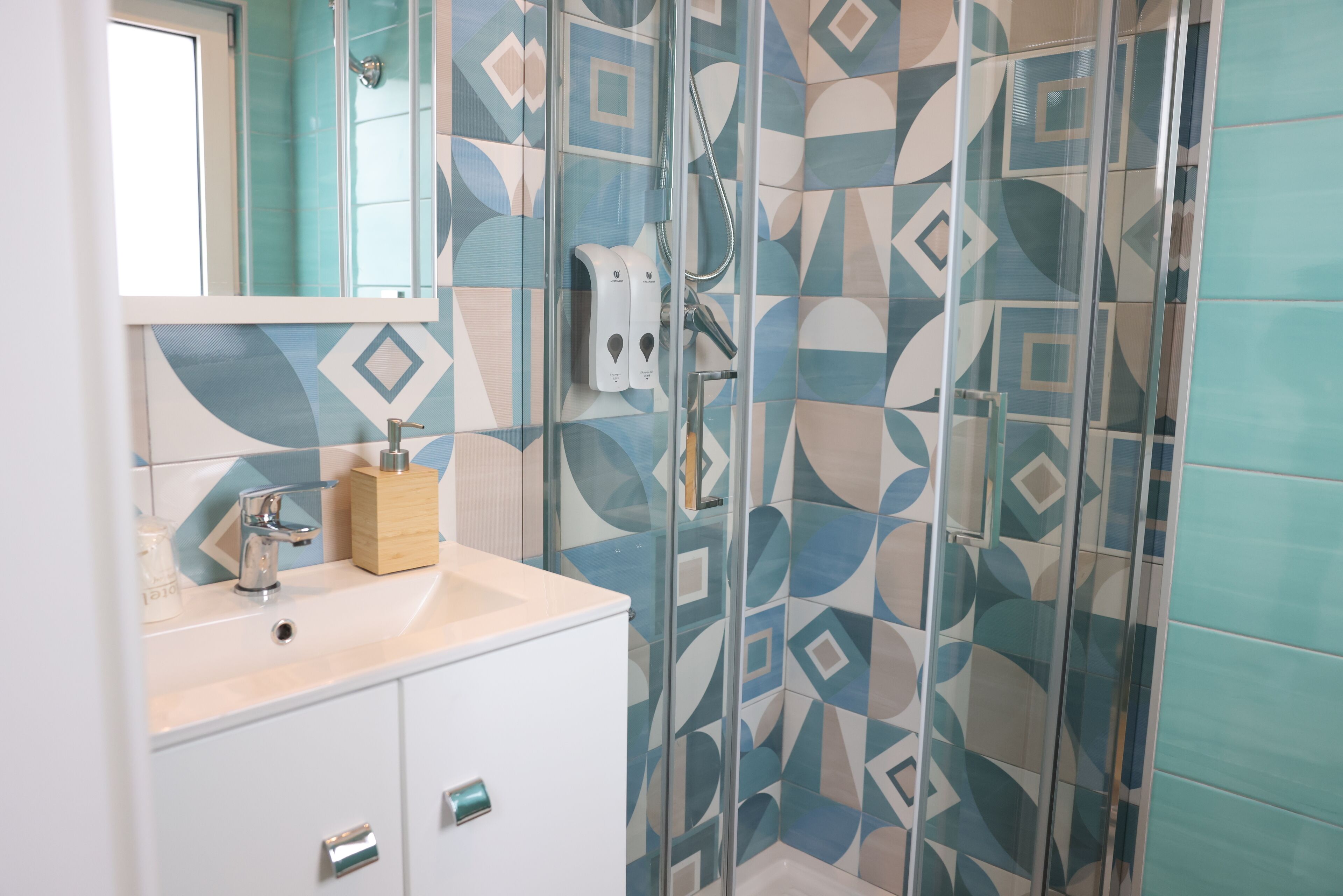 A bathroom with a geometric tile pattern, a glass shower enclosure, a white sink with a wooden soap dispenser, and a mirror above the sink.