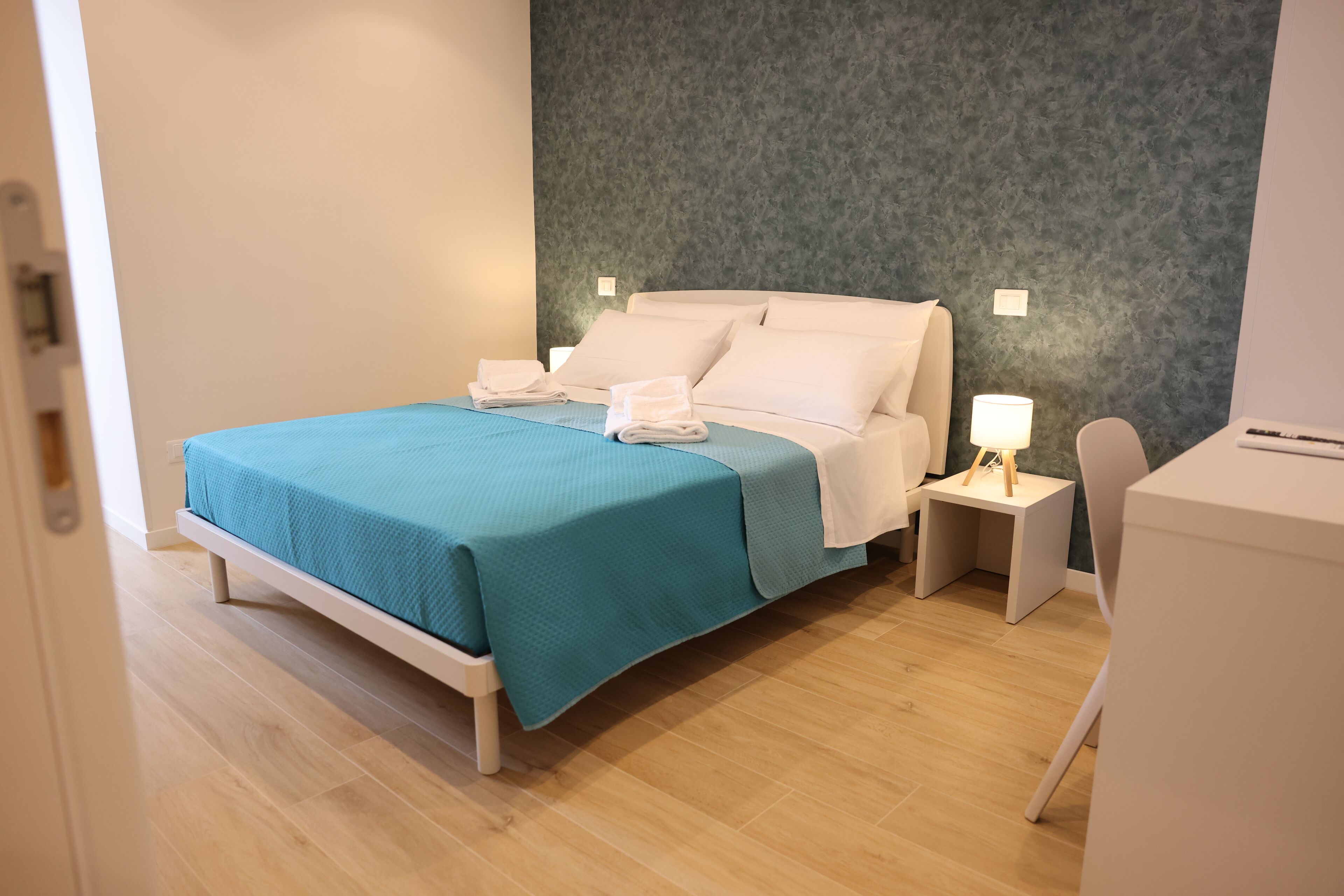 A neatly made bed with a teal bedspread and white pillows, a bedside table with a lamp, and a white chair in a modern room with wooden flooring and a textured gray wall.
