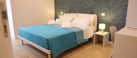 Un letto rifatto con una coperta color petrolio e cuscini bianchi, un comodino con una lampada e una sedia bianca in una stanza moderna con pavimento in legno e parete grigia con texture.