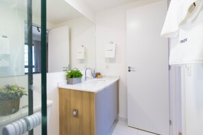 Un baño moderno con un mueble de tocador de madera, un lavabo y un espejo. Hay una planta en maceta sobre el mueble y una toalla colgada en la puerta.