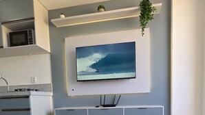 A modern hotel room with a large bed, a flat-screen TV mounted on the wall, a small kitchen area, and a potted plant.
