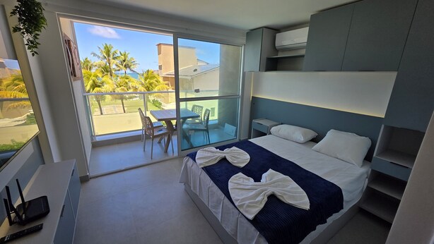 A modern hotel room with a bed, a desk, and a balcony with a view of palm trees and a building.