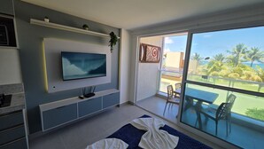 A modern living room with a flat-screen TV mounted on the wall, a balcony with a glass door, and a view of palm trees and the ocean.
