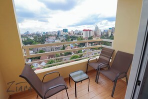 A balcony with two chairs and a small table, overlooking a cityscape with buildings and greenery.