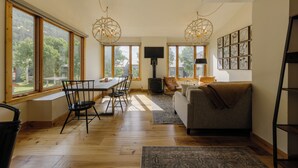 A spacious living area with a dining table, chairs, a sofa, and a fireplace. Large windows provide natural light and a view of the outdoors. - Cozy Mountain Condo by InvitedHome | In Town, Walkable to Ski / Shops (Telluride)