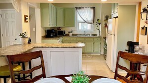 Uma cozinha compacta com armários verdes, uma ilha central e um espaço de refeições com cadeiras de madeira e uma mesa posta para dois.