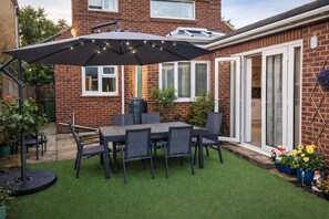 Une terrasse avec une table et des chaises, un grand parasol et une maison en briques dotée d’une porte-fenêtre coulissante.