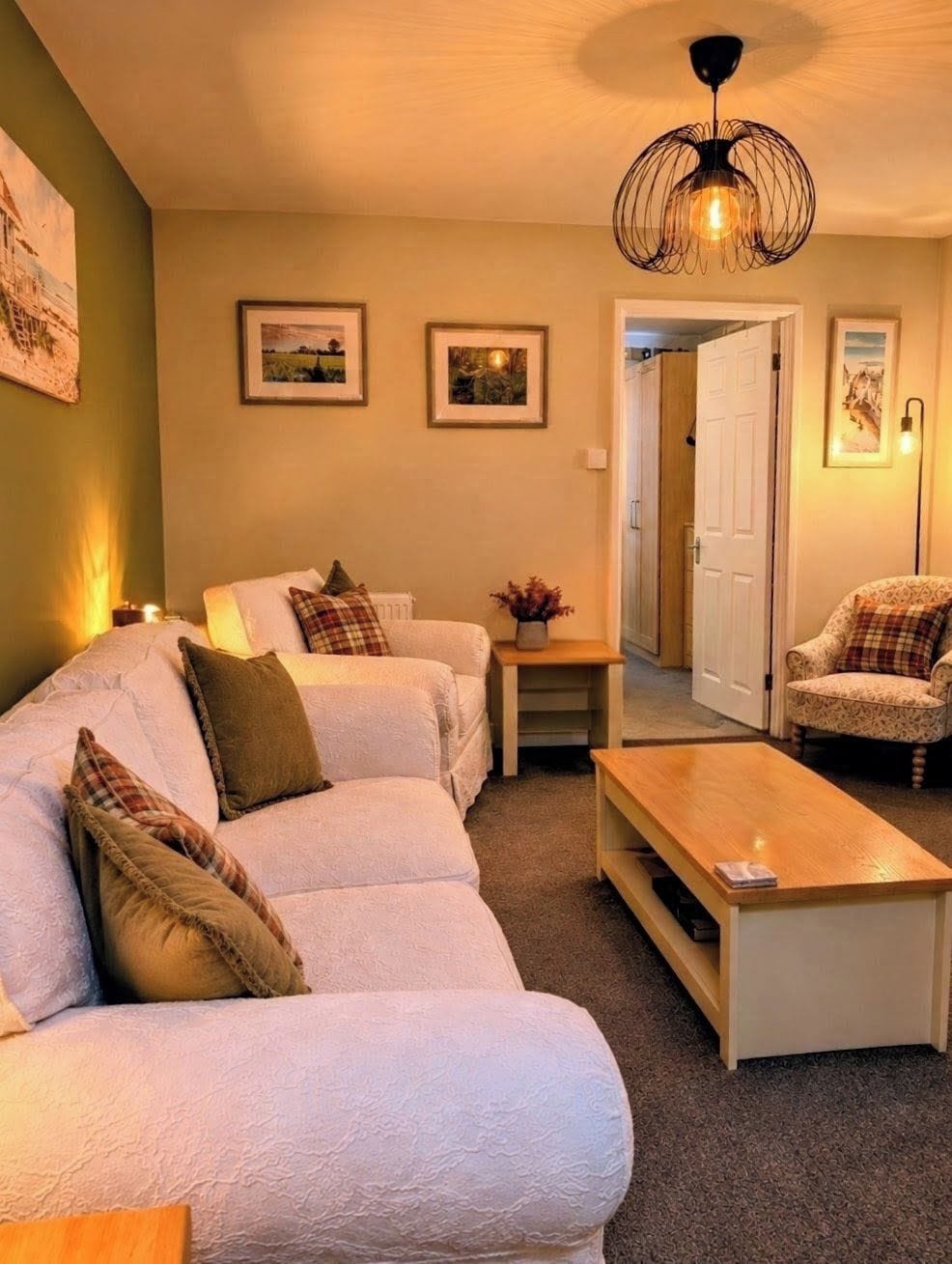 A cozy living room with a sofa, a coffee table, and a patterned armchair. There are framed pictures on the wall and a unique ceiling light fixture.