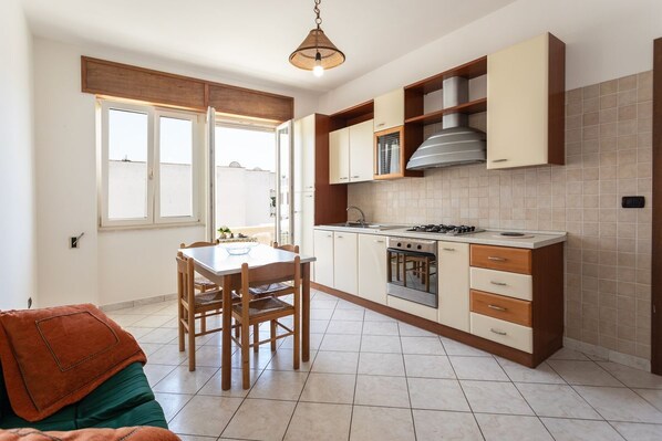 A modern kitchen with white cabinets, a gas stove, and a built-in oven. There is a dining table with chairs and a window with blinds.