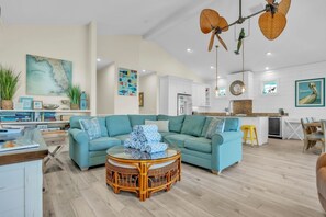 Een ruime woonkamer met een blauwgroene bank, een rond rieten bijzettafeltje, en een keuken op de achtergrond.