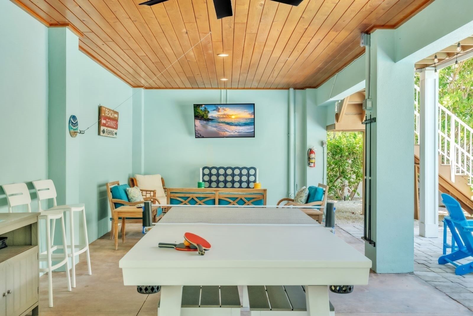 A room with a ping pong table, a sofa with cushions, a wall-mounted TV, and a sign indicating a beach location.