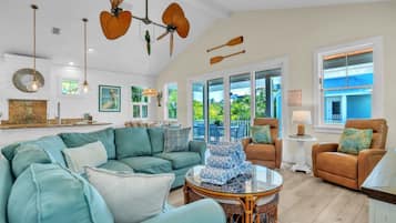 A spacious living room with a large sofa, a coffee table, and a dining area with chairs. There are ceiling fans, a view of the outdoors, and a large window.