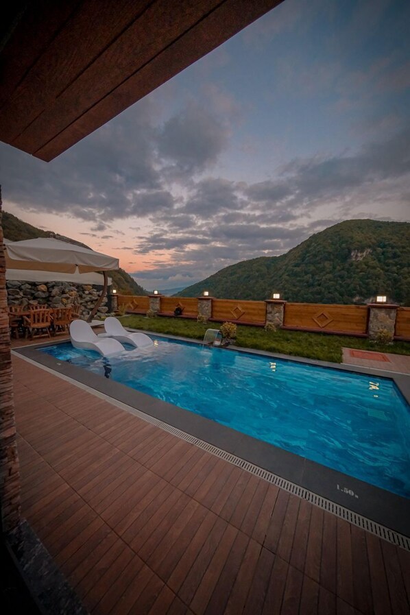 Ein Swimmingpool mit Liegestühlen und einer Holzterrasse, umgeben von einer Steinmauer und Blick auf einen Berg.