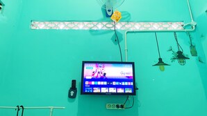 Una habitación con paredes color turquesa, un televisor montado mostrando un juego, un ventilador de pared y una regleta de enchufes.