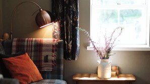 A cozy living room with a plaid sofa, a floor lamp, a window with curtains, and a small table with a vase of flowers. - Charming 1-bedroom cottage - the perfect couples escape! (Mossdale, Castle Douglas)