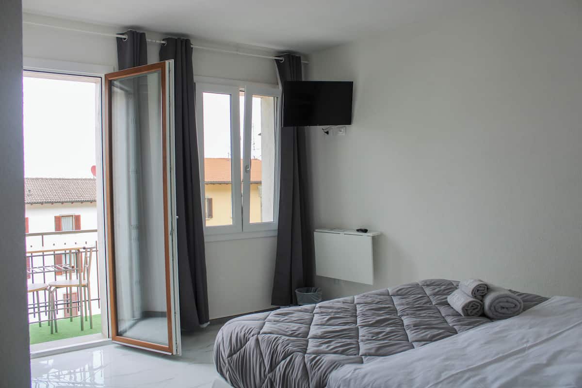 Une chambre avec un lit, un téléviseur fixé au mur, une porte-fenêtre coulissante donnant sur un balcon, et une petite table avec des chaises à l’extérieur.