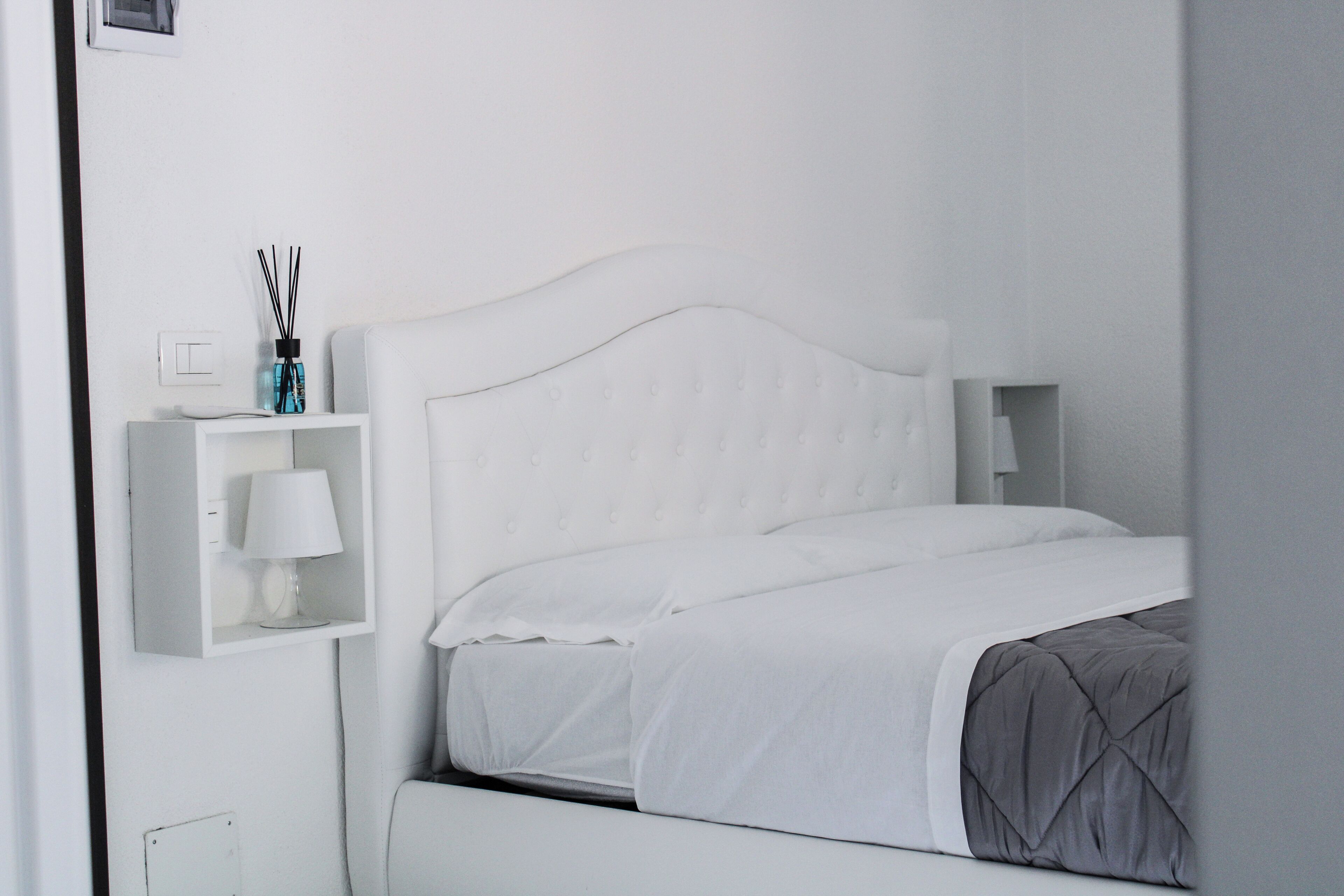 Ein weißes Bett mit einer grauen Decke, ein weißer Nachttisch mit einer Lampe und ein kleines Regal mit einer Lampe und einer Wasserflasche.