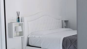 Ein weißes Bett mit einer grauen Decke, ein weißer Nachttisch mit einer Lampe und ein kleines Regal mit einer Lampe und einer Wasserflasche.
