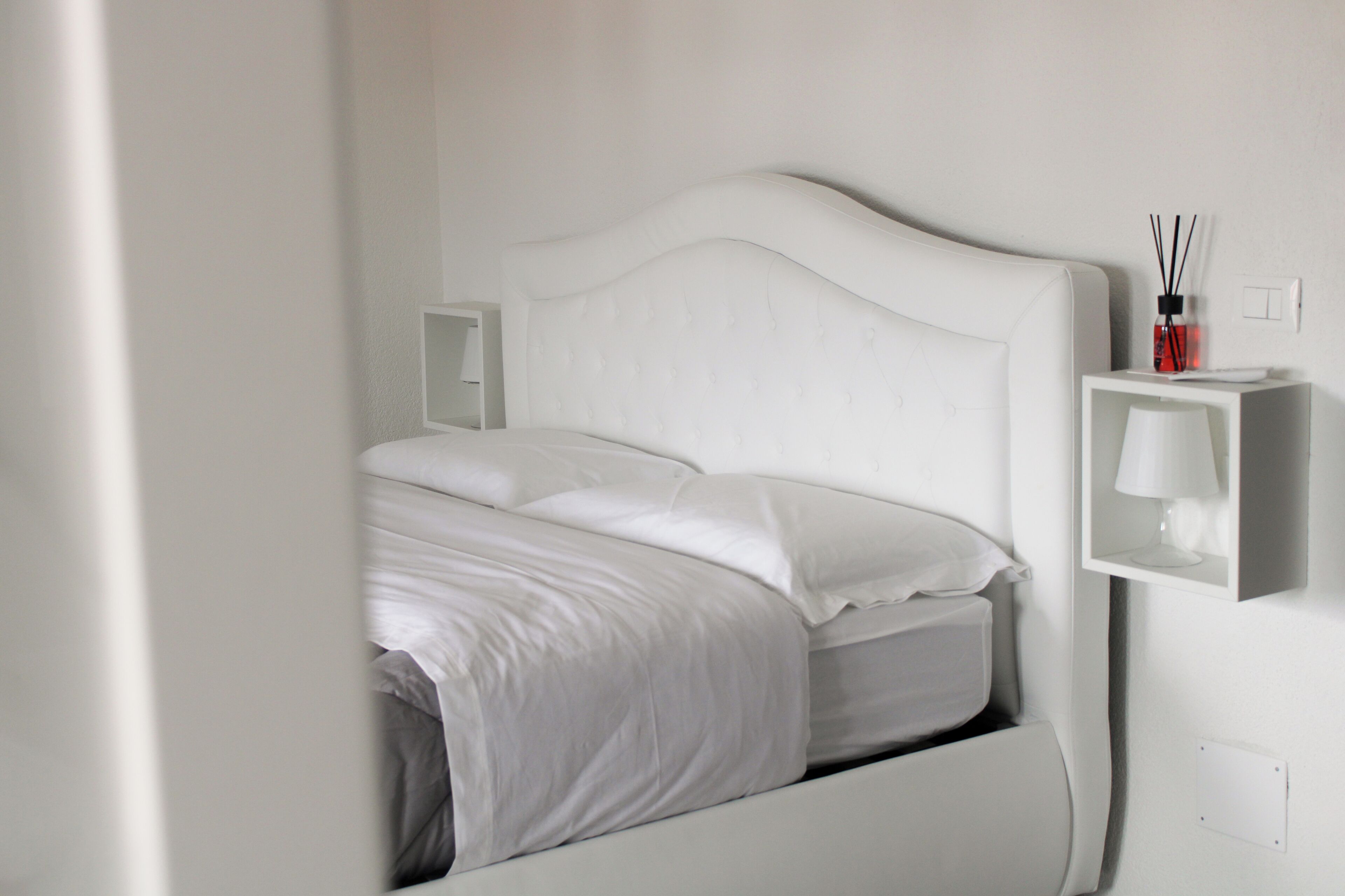 Un lit blanc avec une tête de lit, une table de chevet avec une lampe et une petite étagère sur laquelle se trouvent un flacon et un diffuseur à tiges.