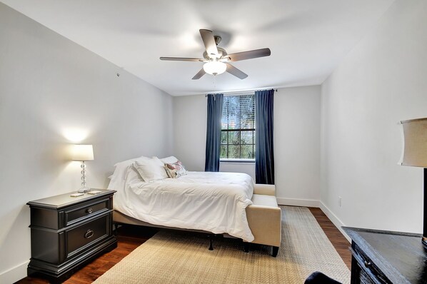 Un dormitorio con cama, mesita de noche, ventilador de techo, ventana con cortinas y vistas al exterior.