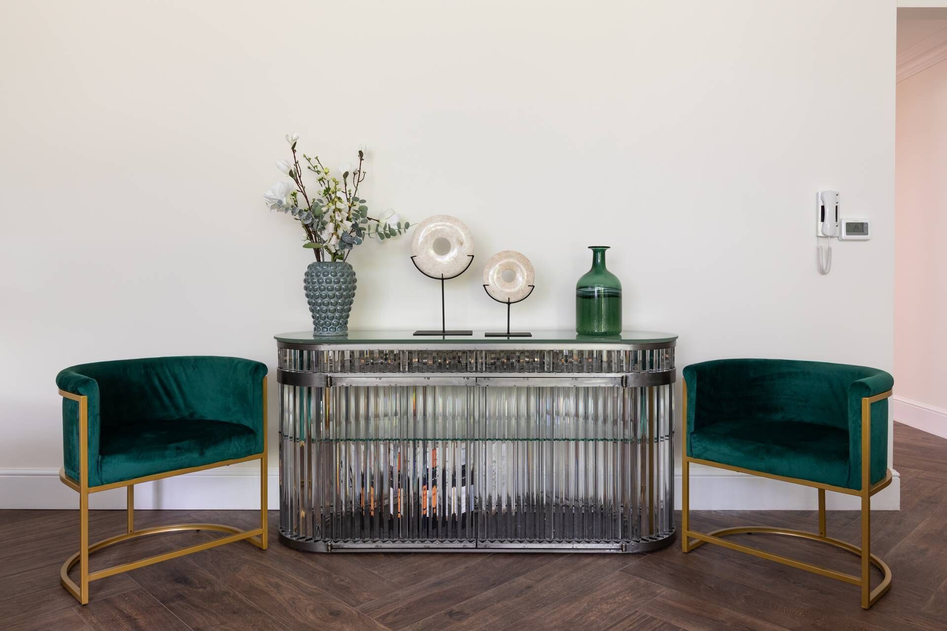 A room with a glass-topped console table, two green velvet armchairs, and a vase with white flowers.