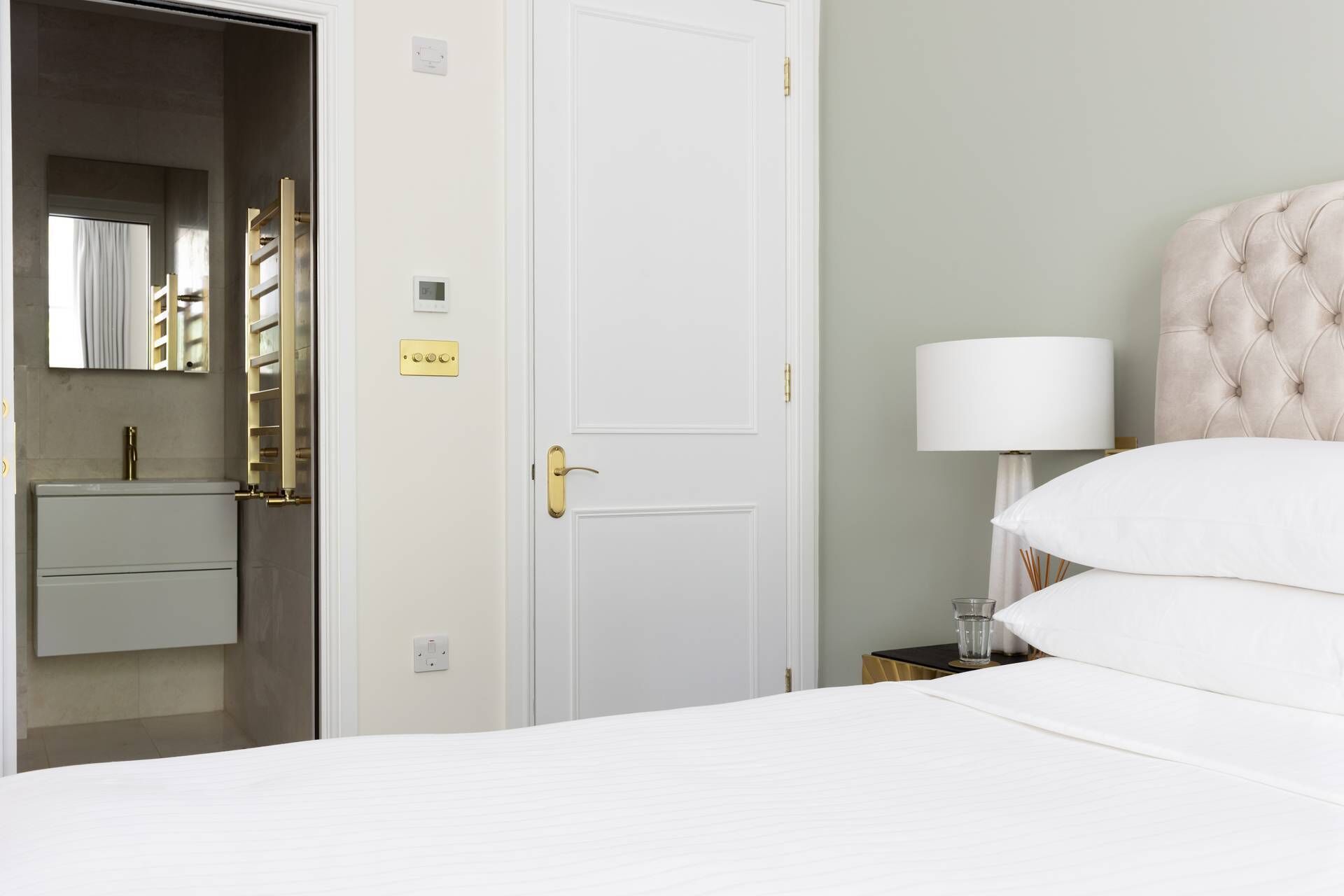 Een hotelkamer met een bed, nachtkastje met lamp, dressoir en een deur met spiegel en lichtschakelaar.