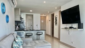 A modern living room with a grey sectional sofa, a kitchen area with a built-in stove, and a mounted flat-screen TV.