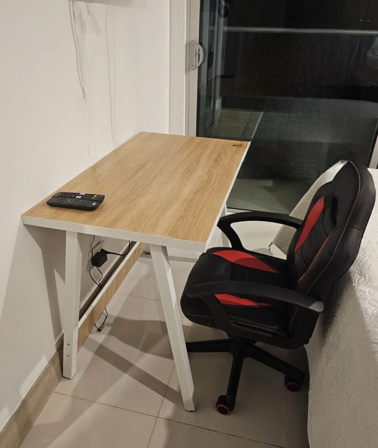 A small office space with a wooden desk, a black and red office chair, and a glass door leading to a balcony.