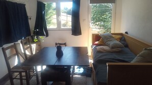 A small, cozy room with a wooden table, a single chair, a bed with pillows, and a window with curtains.