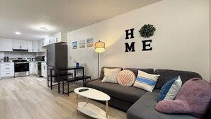 Una sala de estar moderna con un sofá gris, una mesita blanca y una cocina al fondo.