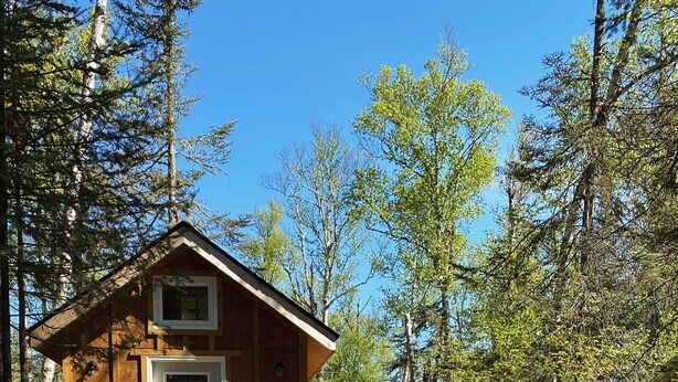 A wooden cabin with a deck, two Adirondack chairs, and a metal fire pit.