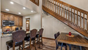 Eine Küche mit Holzausstattung, einer Mikrowelle und einer Kaffeemaschine. Es gibt einen Essbereich mit einem Holztisch und Stühlen. Im Hintergrund ist eine Treppe mit Holzwurf sichtbar.