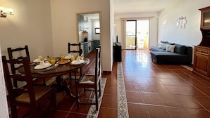 Comedor con una mesa redonda, sillas y productos para el desayuno. Se observa una cocina al fondo con armarios y un lavamanos. En primer plano, un salón con un sofá y un televisor.