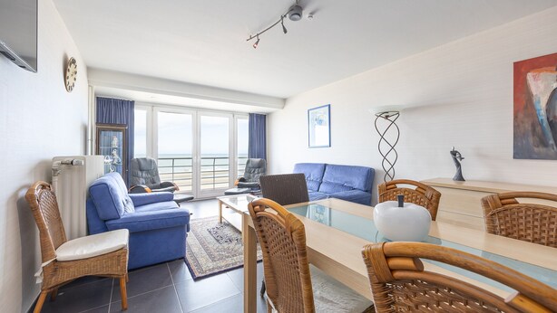 Una sala de estar moderna con un sofá azul, sillas de mimbre, una mesa de comedor de vidrio y vistas al mar a través de amplios ventanales.