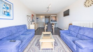 Una sala de estar con dos sofás azules, una mesa de centro y un tapete estampado. Hay un reloj en la pared y un televisor montado sobre el sofá.