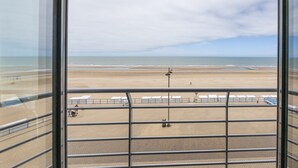 Vista de una playa desde un balcón con barandillas de metal, arena dorada y mar tranquilo.
